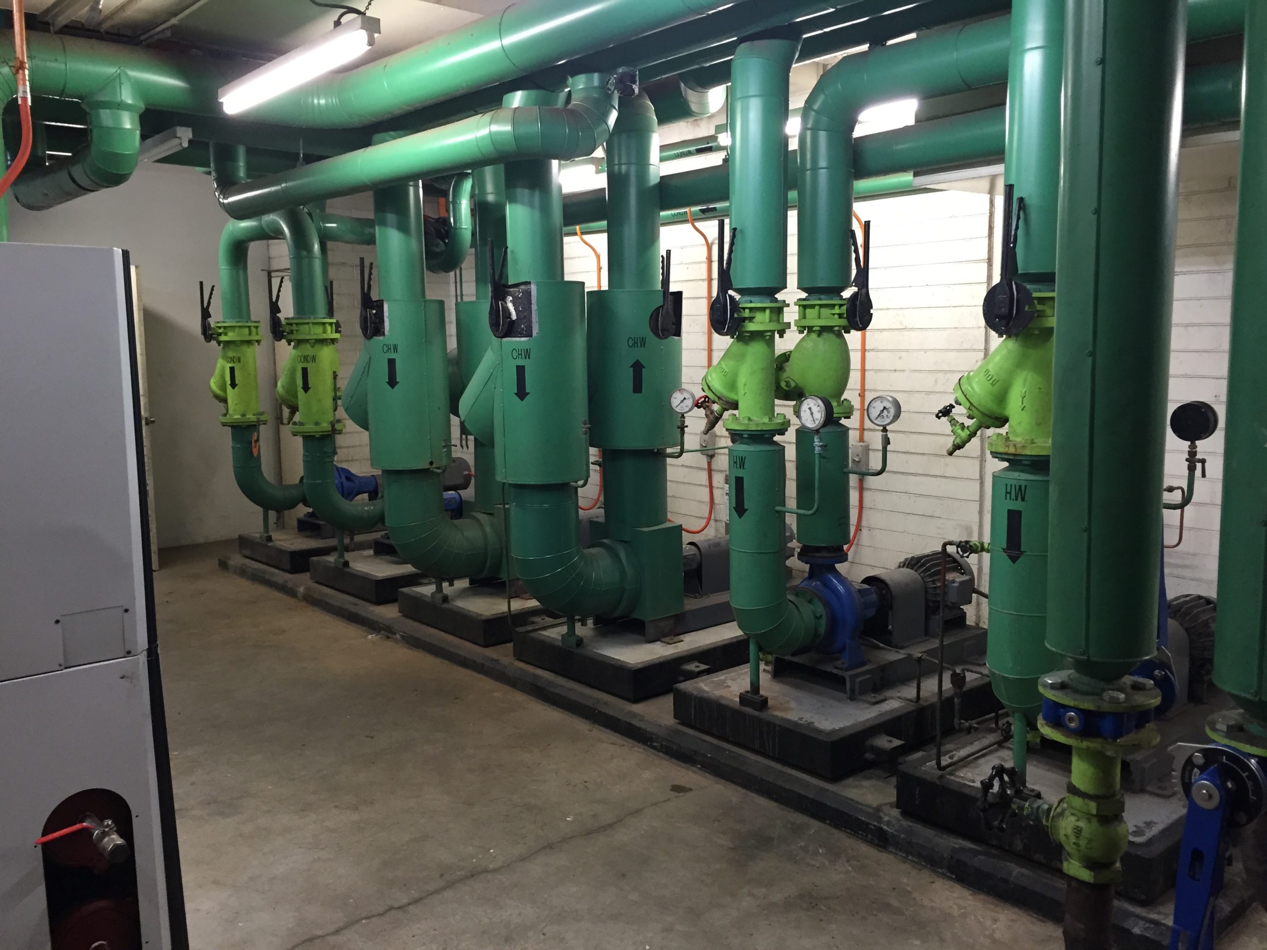 Industrial mechanical room with multiple green pipes, valves, and pumps connected to HVAC equipment.