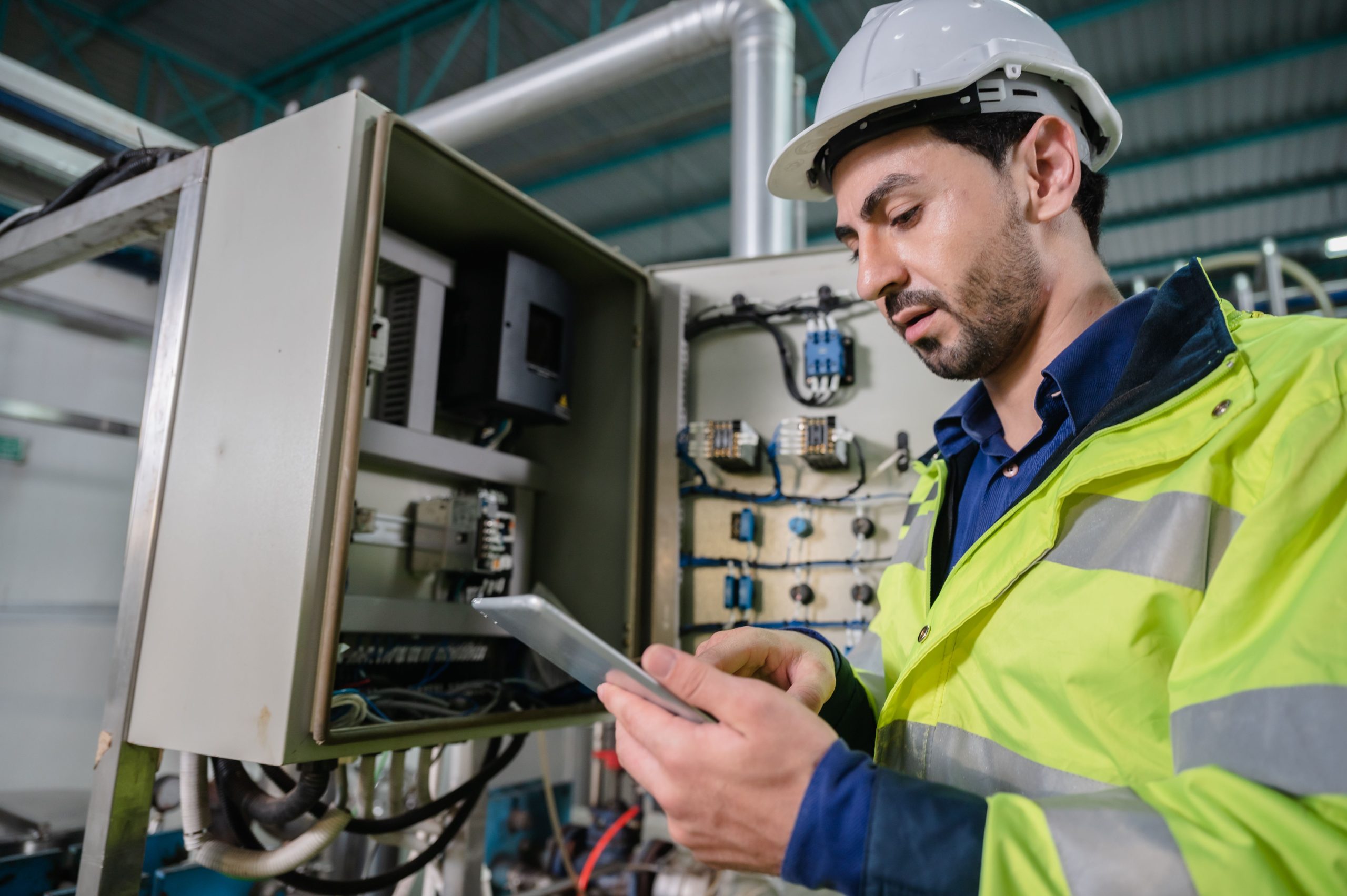 Technician in safety gear using a tablet while inspecting an open electrical control panel.